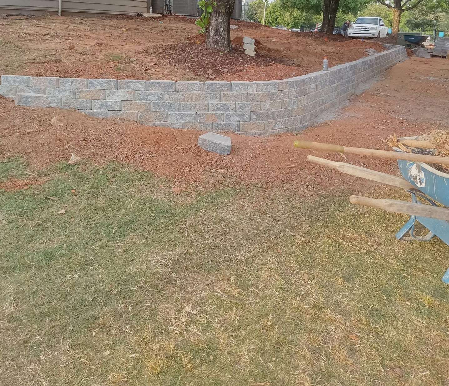 Curved gray block retaining wall during installation with exposed soil and tools nearby