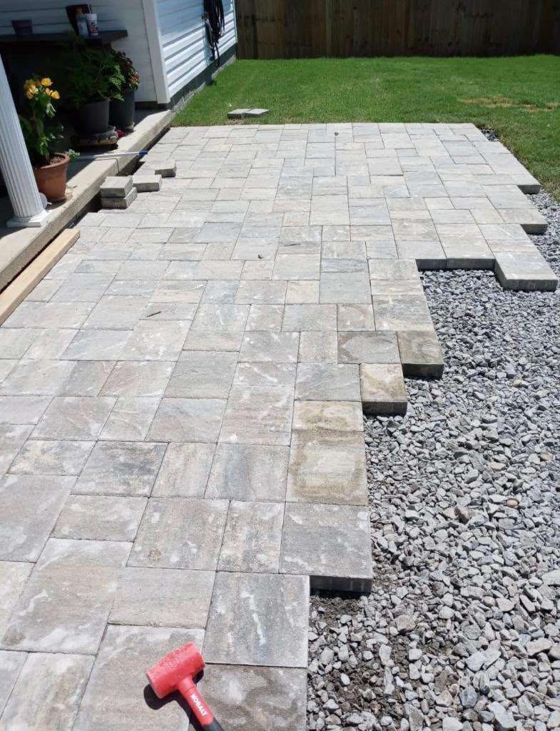 Paver patio installation beside a porch with an unfinished edge and aggregate base